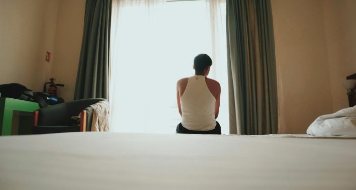 A man sits indoors facing a bright window, lost in thought, casting an introspective mood.