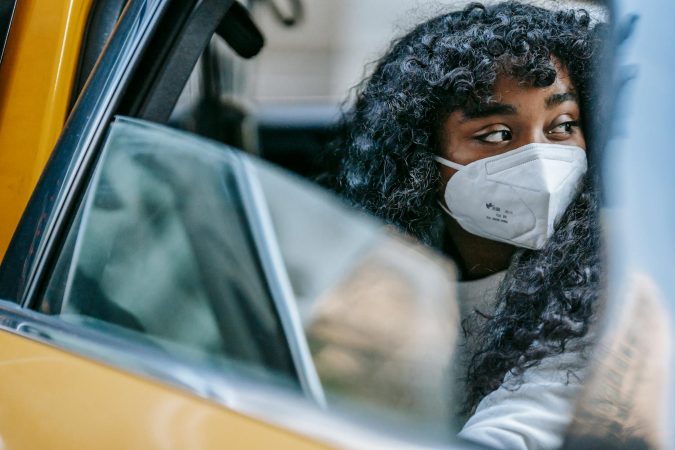Black young lady in casual clothes and protective mask sitting in yellow cab with open door and looking away thoughtfully in daytime in city street