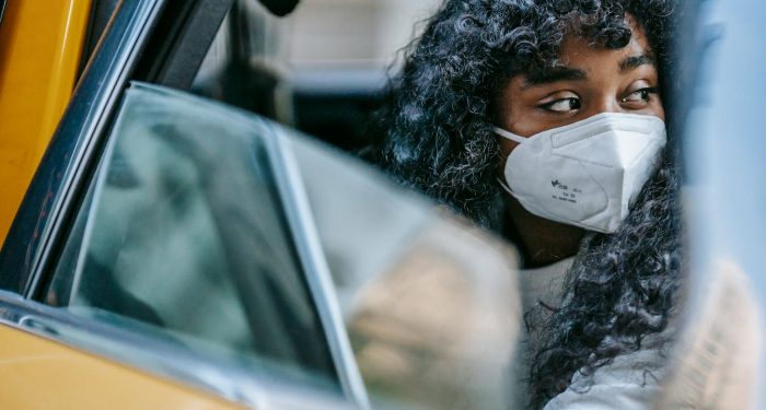 Black young lady in casual clothes and protective mask sitting in yellow cab with open door and looking away thoughtfully in daytime in city street