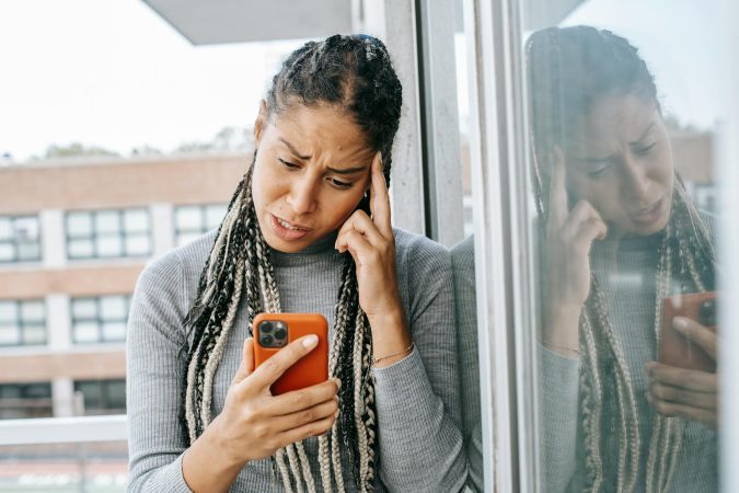 Unhappy woman with braids looking at smartphone on balcony, expressing dissatisfaction.