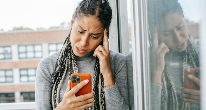 Unhappy woman with braids looking at smartphone on balcony, expressing dissatisfaction.