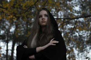 Thoughtful young woman outdoors in a dark hooded cape, with arms crossed, conveying a gothic and pensive mood.