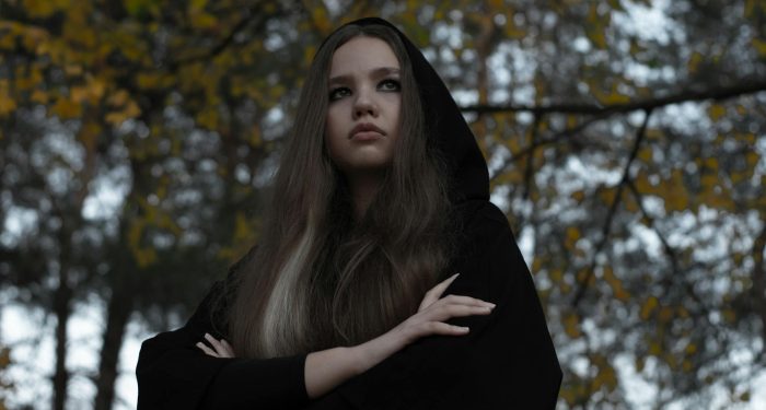 Thoughtful young woman outdoors in a dark hooded cape, with arms crossed, conveying a gothic and pensive mood.