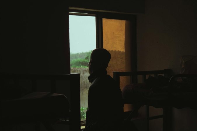 A man in silhouette stands by a window in a dimly lit room with bunk beds, evoking solitude.
