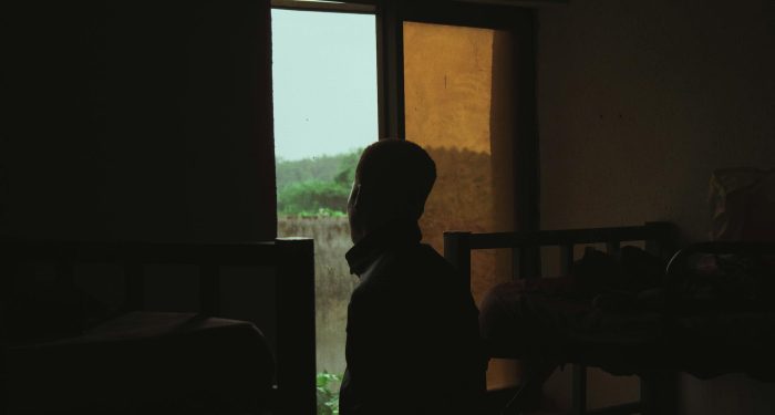 A man in silhouette stands by a window in a dimly lit room with bunk beds, evoking solitude.