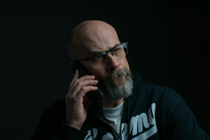 Senior bald man wearing eyeglasses, engages in a phone call against a dark background.