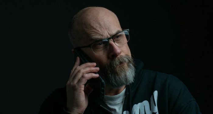 Senior bald man wearing eyeglasses, engages in a phone call against a dark background.