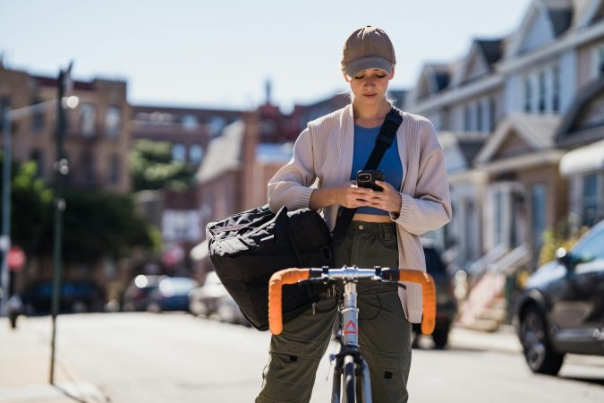 Woman cyclist pauses to check her phone on a sunny city street, balancing urban life and movement.
