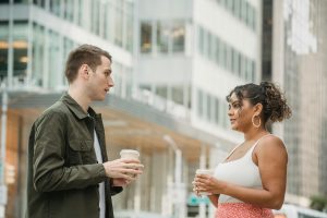 Two young adults enjoying a coffee break outdoors, engaging in conversation amidst an urban setting.