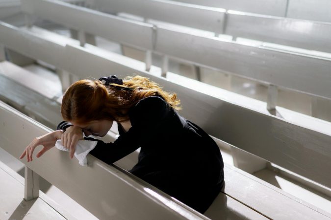 A woman in black clothes leans over pews, grieving alone in an empty church.