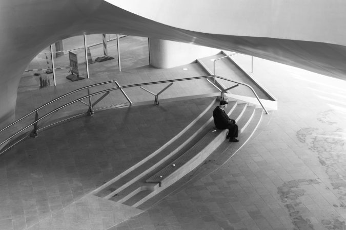 Black and white photo of a lone person seated in a modern architectural space.