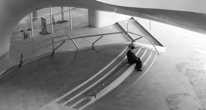 Black and white photo of a lone person seated in a modern architectural space.