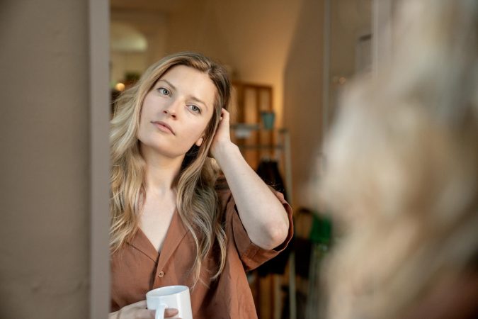 A thoughtful woman holding a coffee cup looks at her reflection in the mirror in a cozy indoor setting.