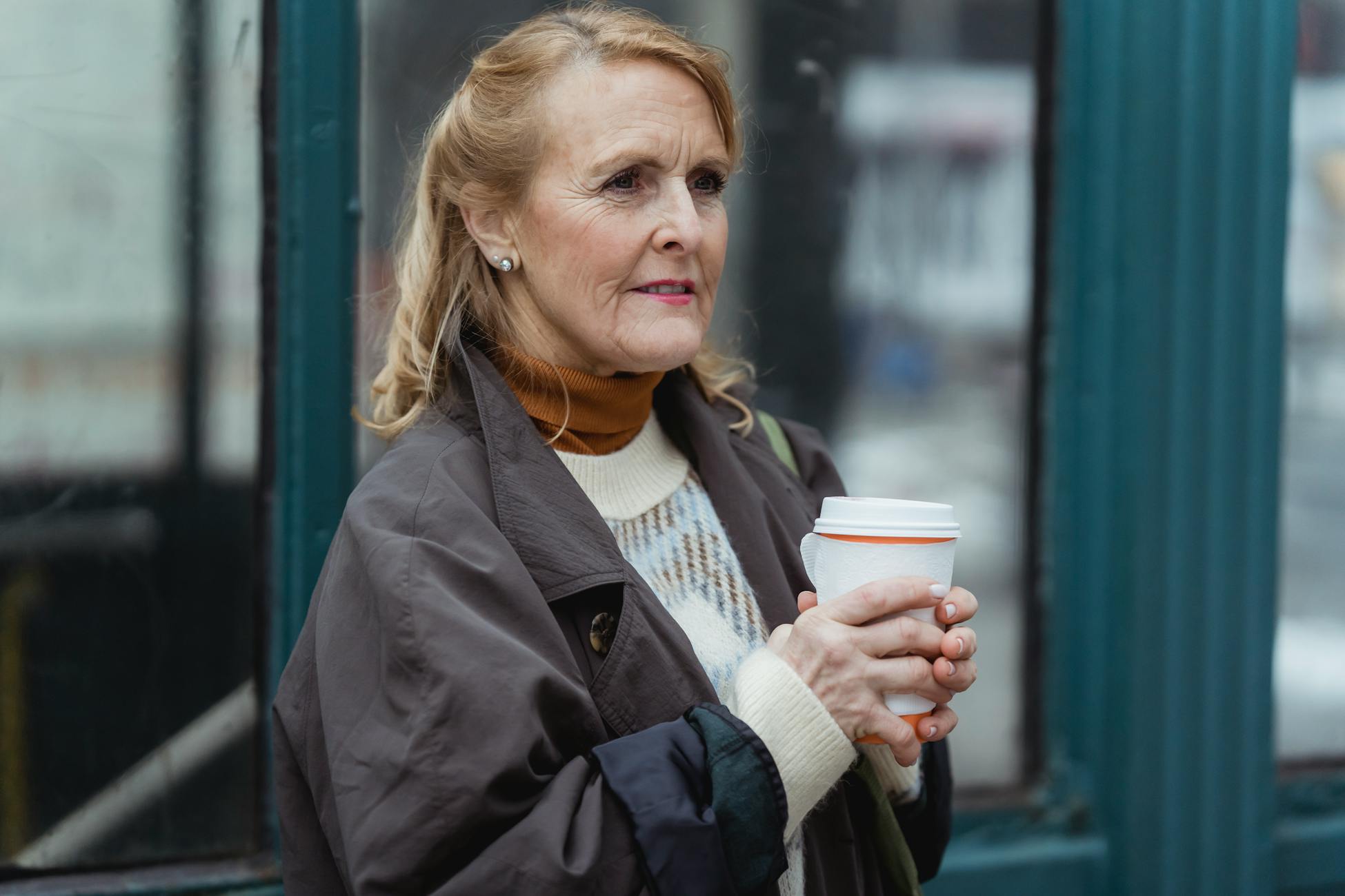 Contemplative elderly female in outerwear with hot beverage to go looking forward in town in daylight