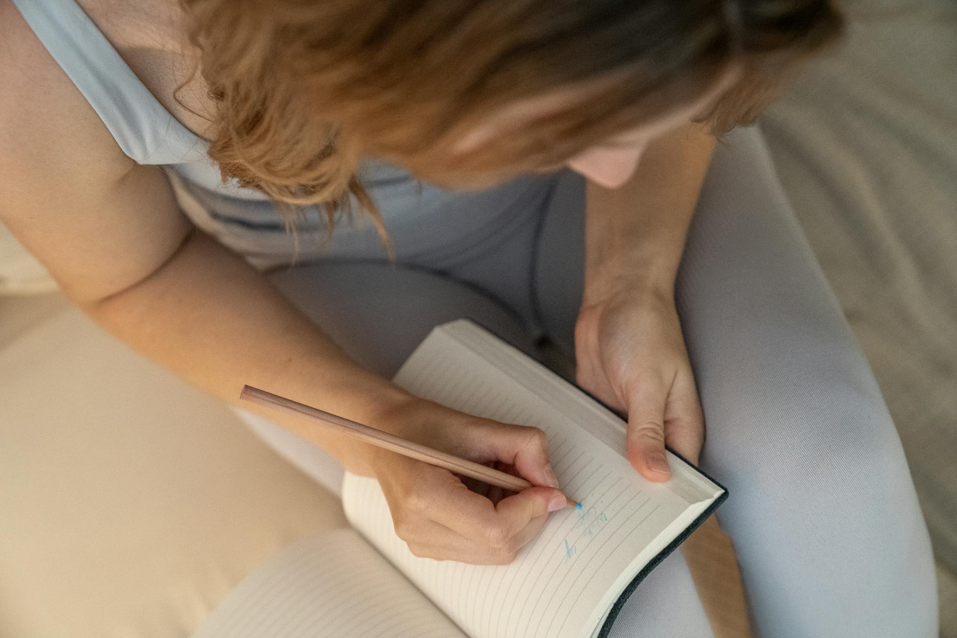 An adult woman writes in a notebook with a pencil, viewed from above.