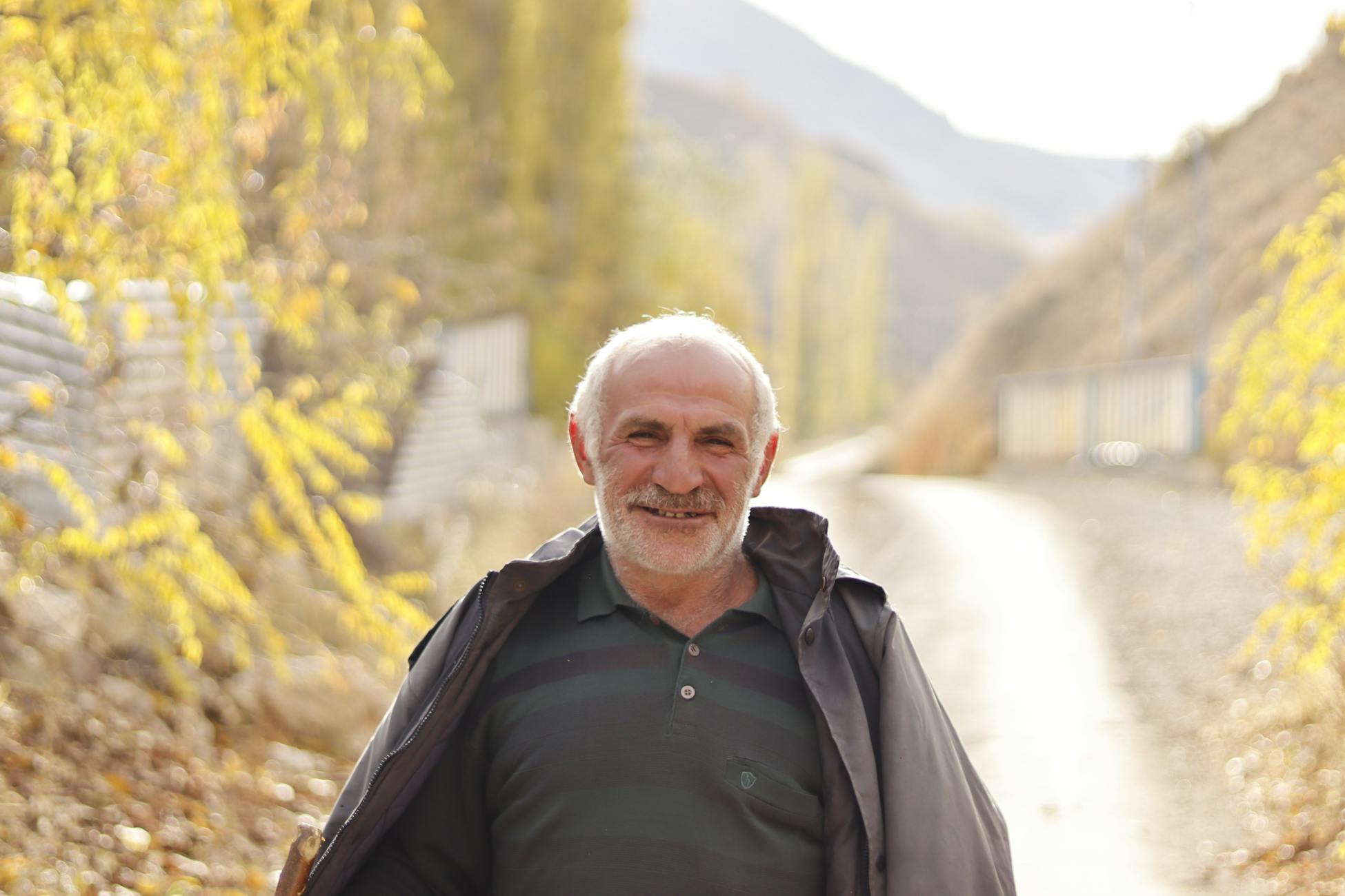 Senior man smiling while standing on a sunlit path in a rural mountain area during fall.