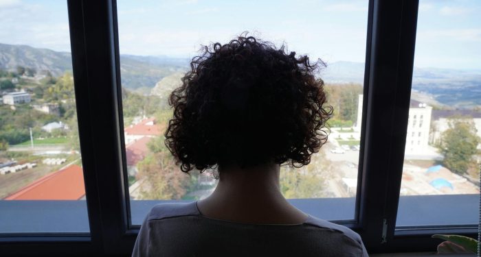 A woman with curly hair gazing out a window at a scenic mountain view, indoors.