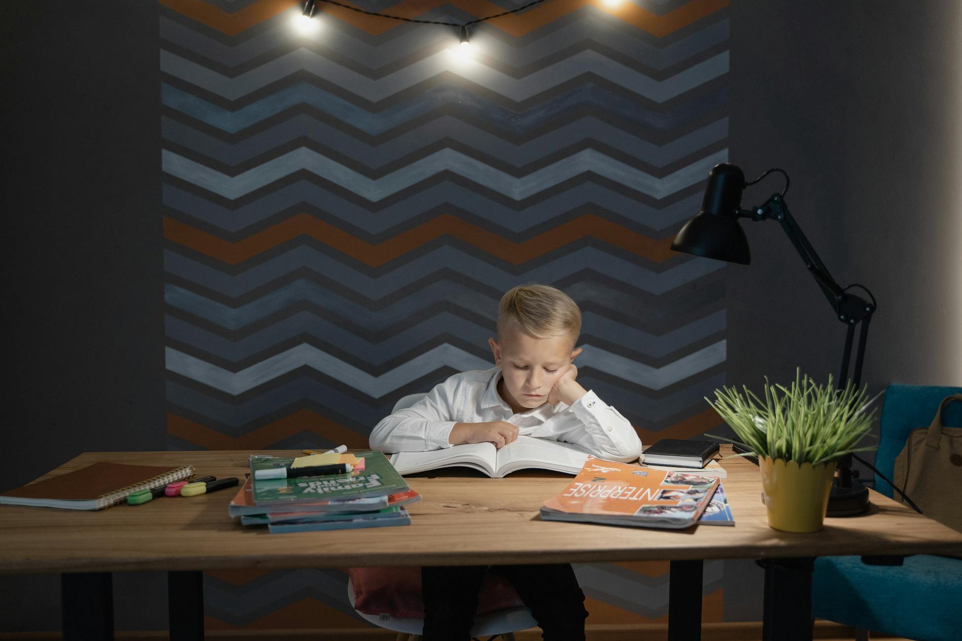 Young boy focused on reading at a desk filled with books and a lamp in dim lighting.