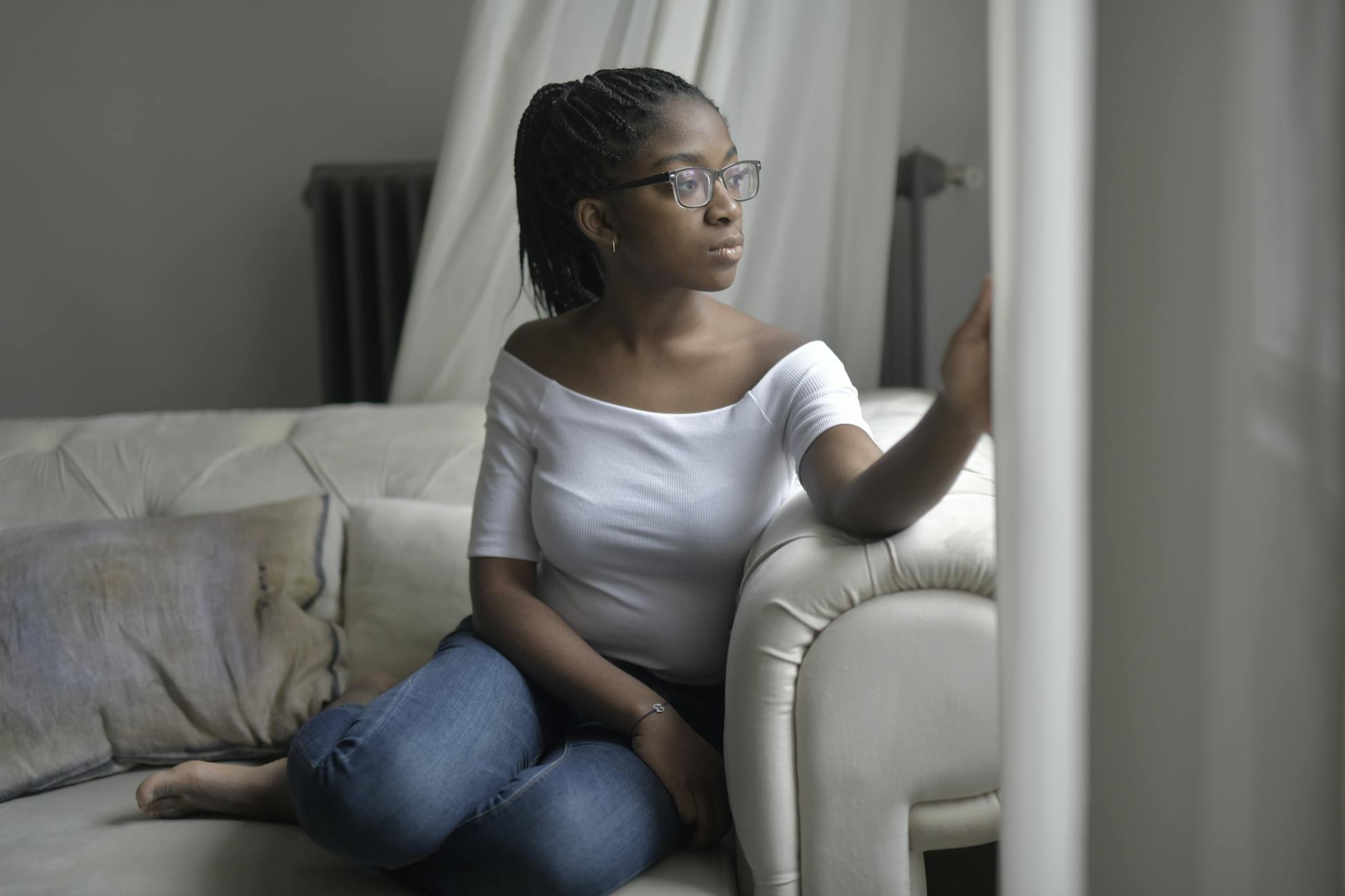 Woman in glasses relaxing on a sofa near window, deep in thought and looking outside.