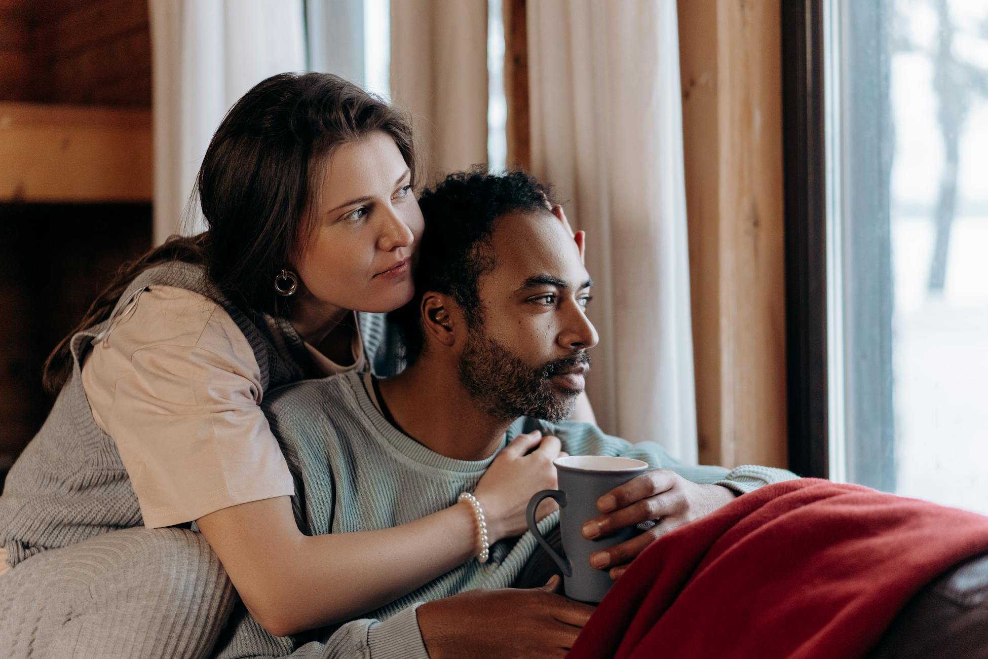 A multiethnic couple enjoys a cozy moment indoors, sitting by a window with a warm embrace.