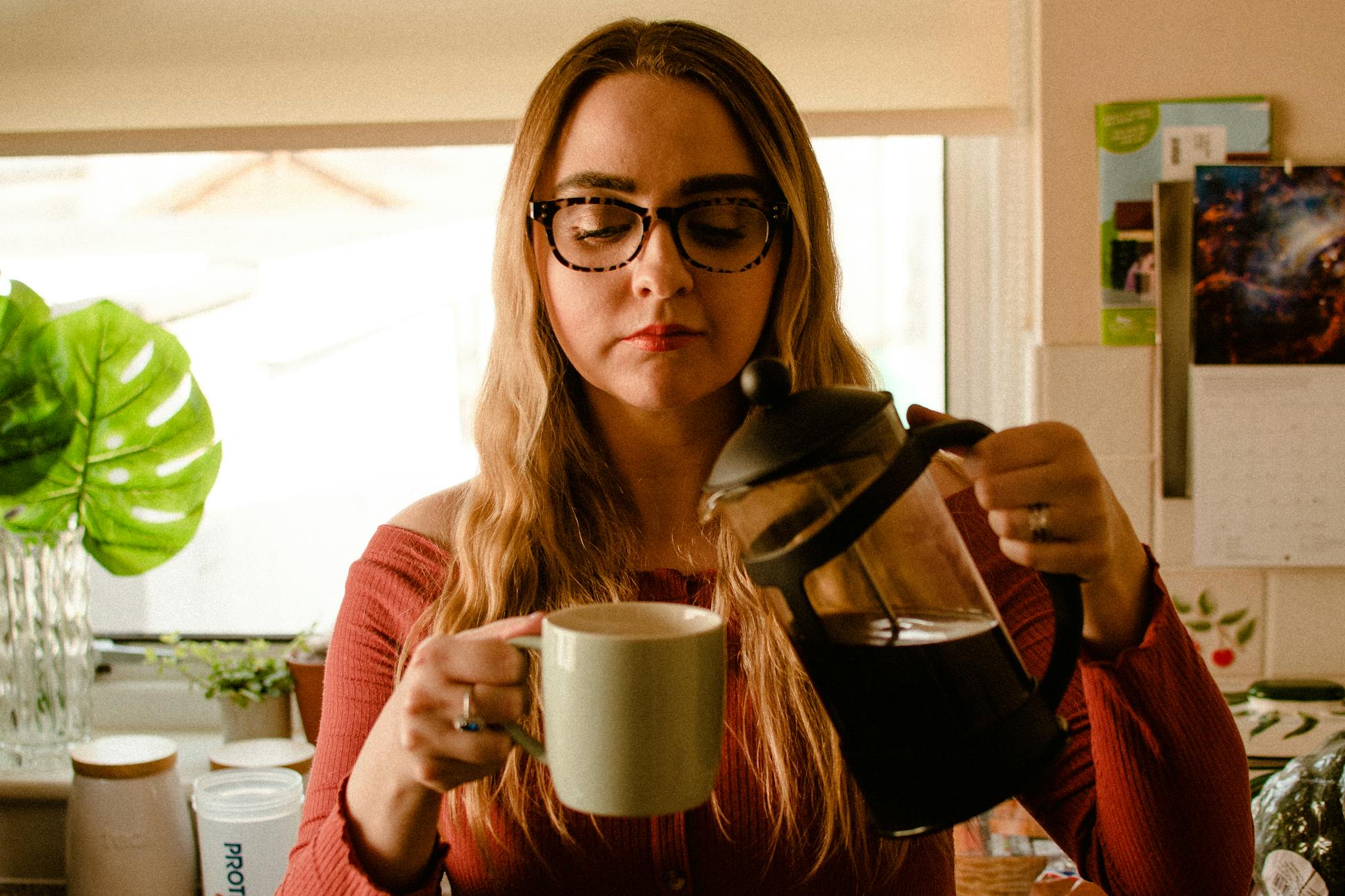 A woman pours coffee into a mug using a French press in a cozy kitchen setting.