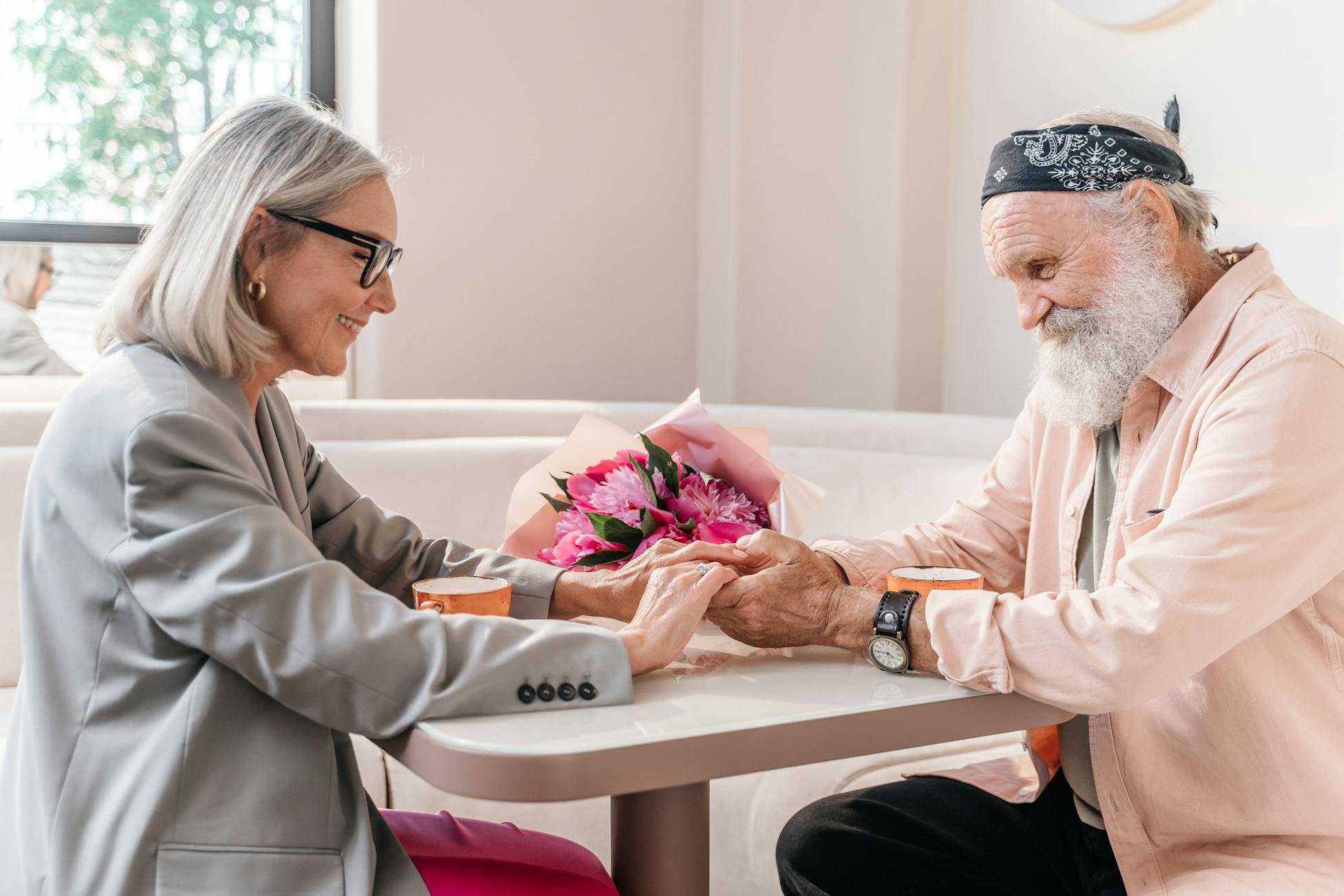 Smiling elderly couple holding hands at a cafe table, surrounded by flowers.