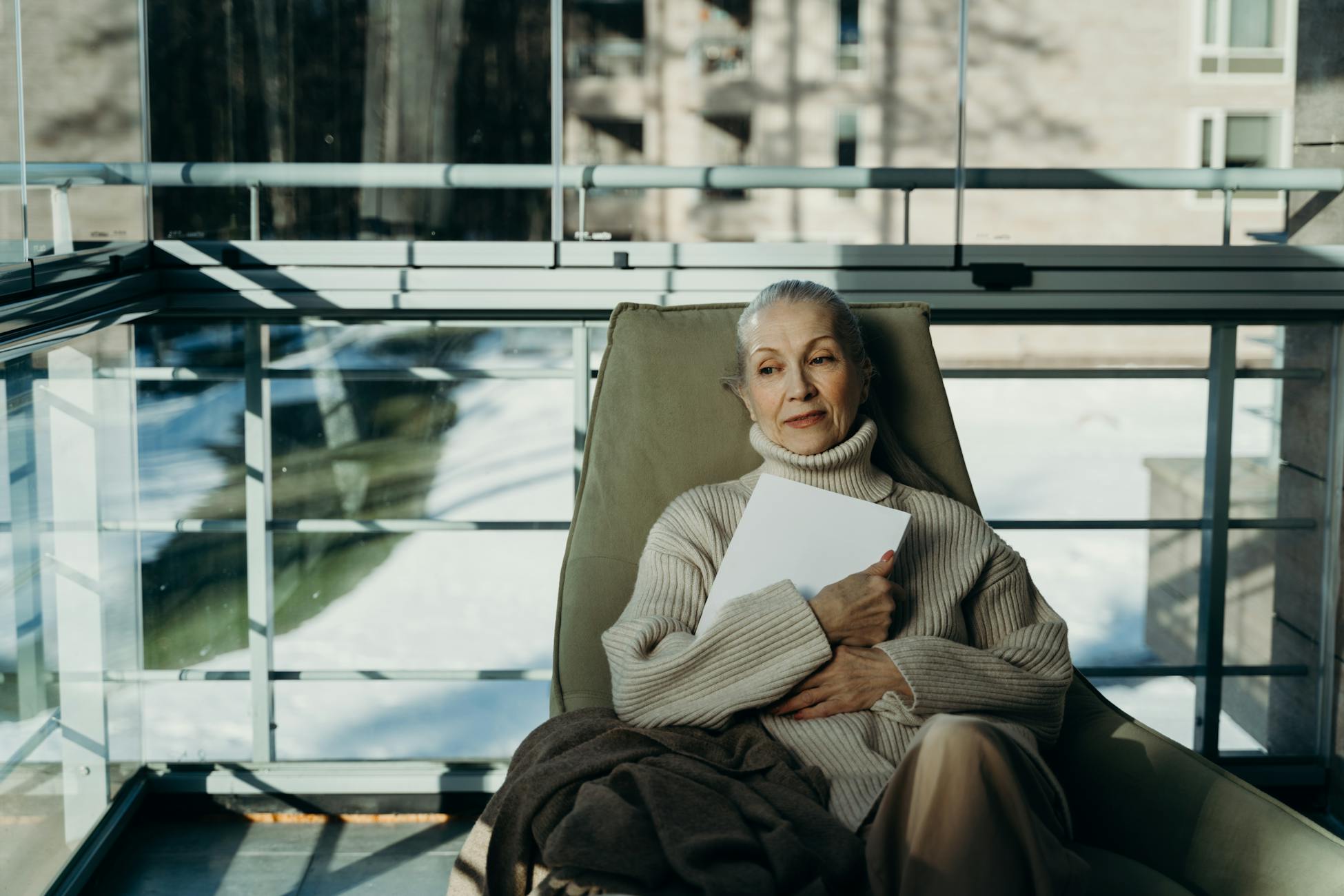 Elderly woman in cozy sweater relaxes indoors with a book in a serene setting.