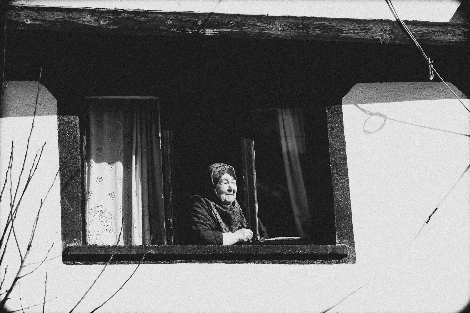 A senior woman with a headscarf looks out from a window in a rural setting, captured in black and white.