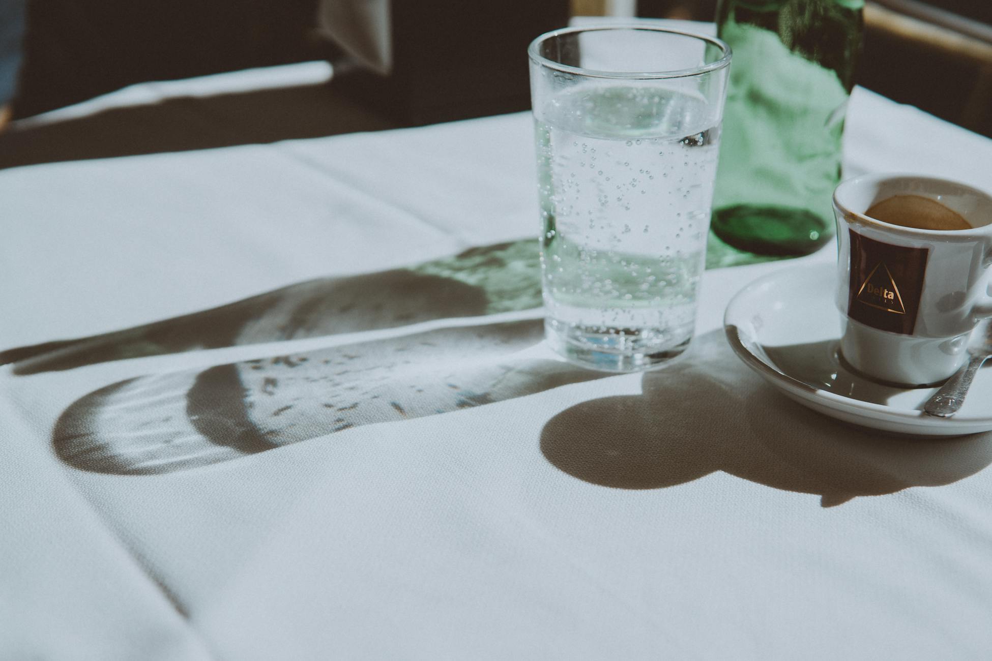 Espresso cup and sparkling water casting shadows on a sunlit table indoors.