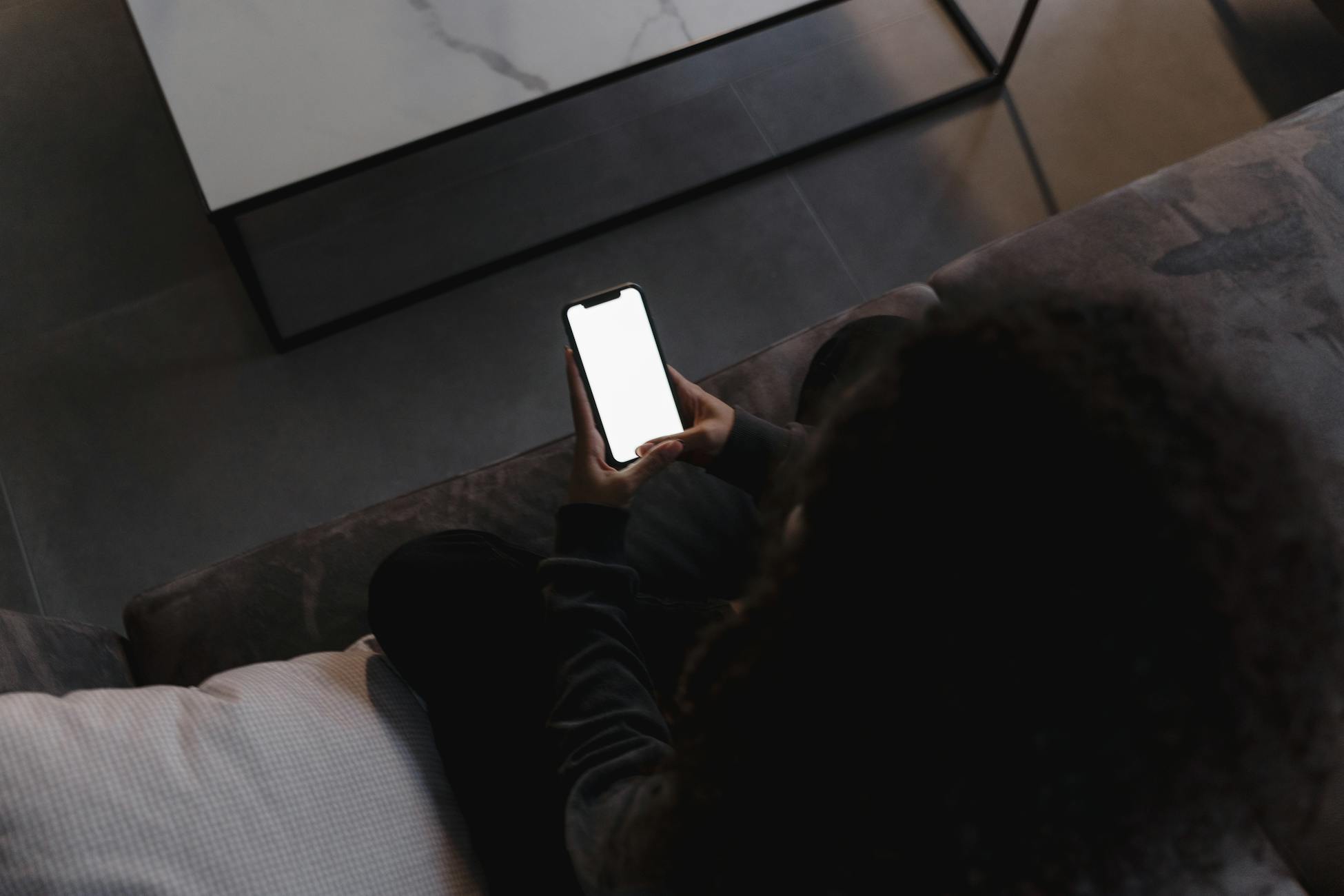 A person holding a smartphone with a blank screen, seated indoors on a couch.