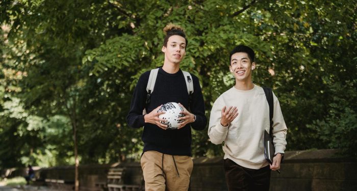 Happy smiling ethnic male friends with backpacks and soccer ball strolling together in lush park and chatting