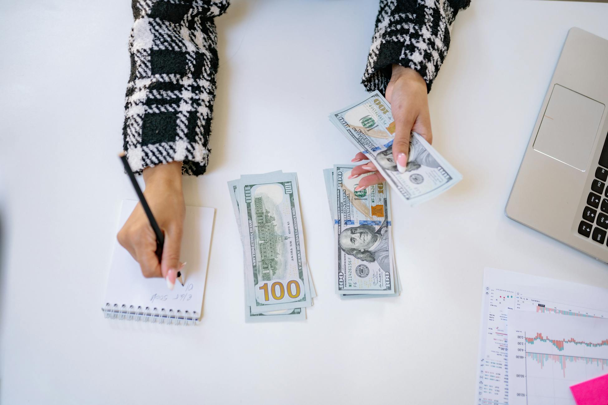 Top view of a person counting US dollars with a notepad at a workspace, indicating financial planning.