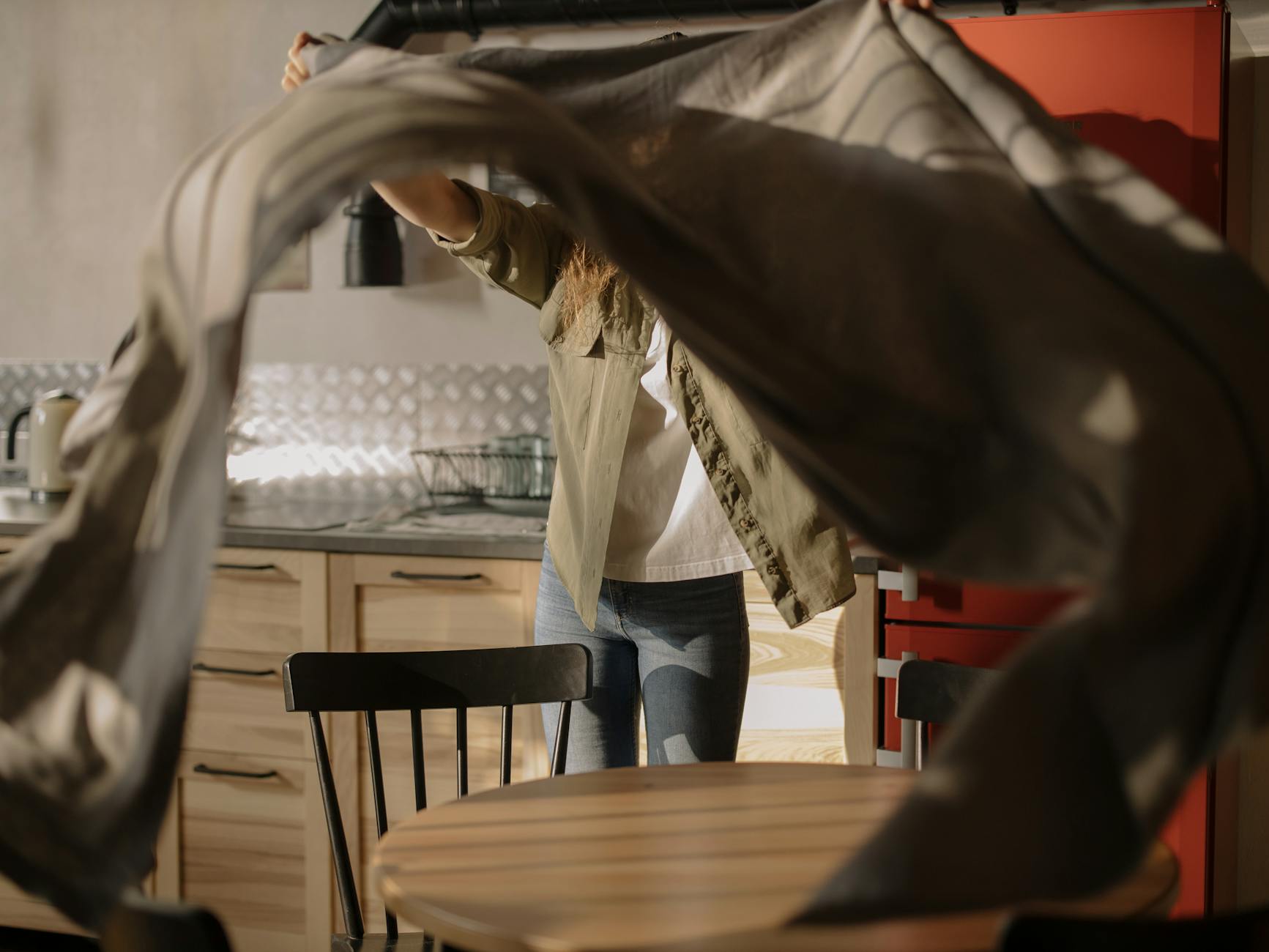 A woman setting a table with a tablecloth in a sunlit kitchen for a cozy home atmosphere.