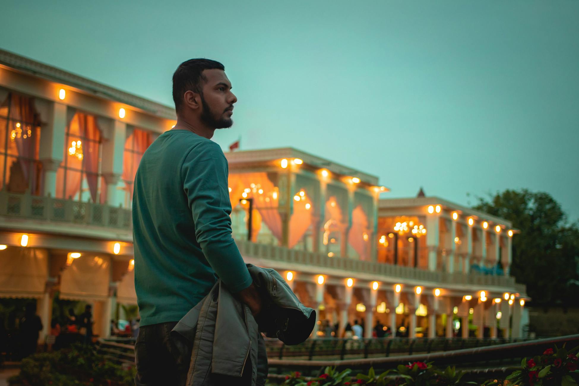 A thoughtful man stands outdoors at twilight, admiring an elegantly lit building, soft lighting.