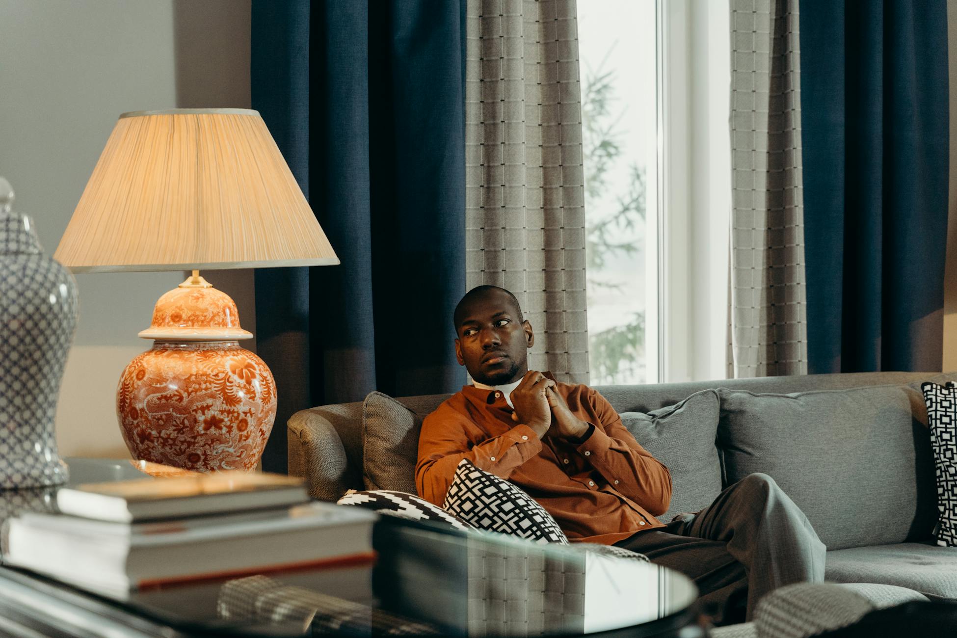 A man in a cozy room sits thoughtfully on a sofa, illuminated by a warm lamp.