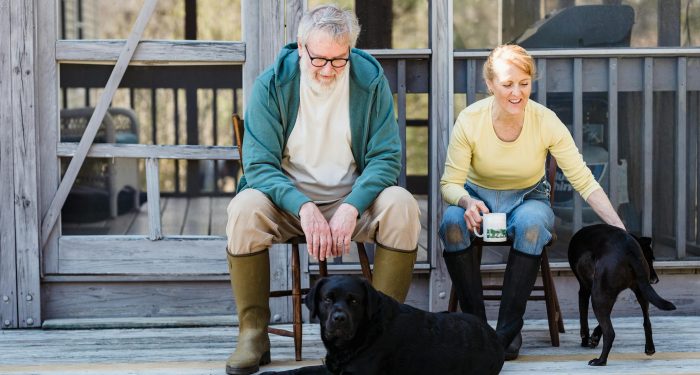 An elderly couple sits with their two dogs on a wooden porch, sharing a peaceful moment outdoors.