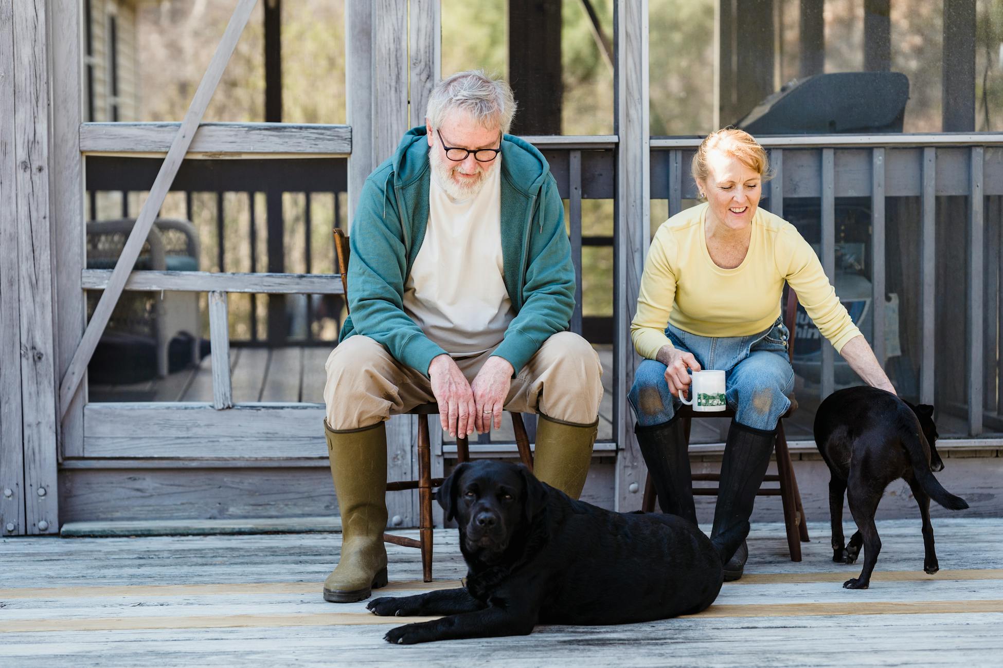 An elderly couple sits with their two dogs on a wooden porch, sharing a peaceful moment outdoors.