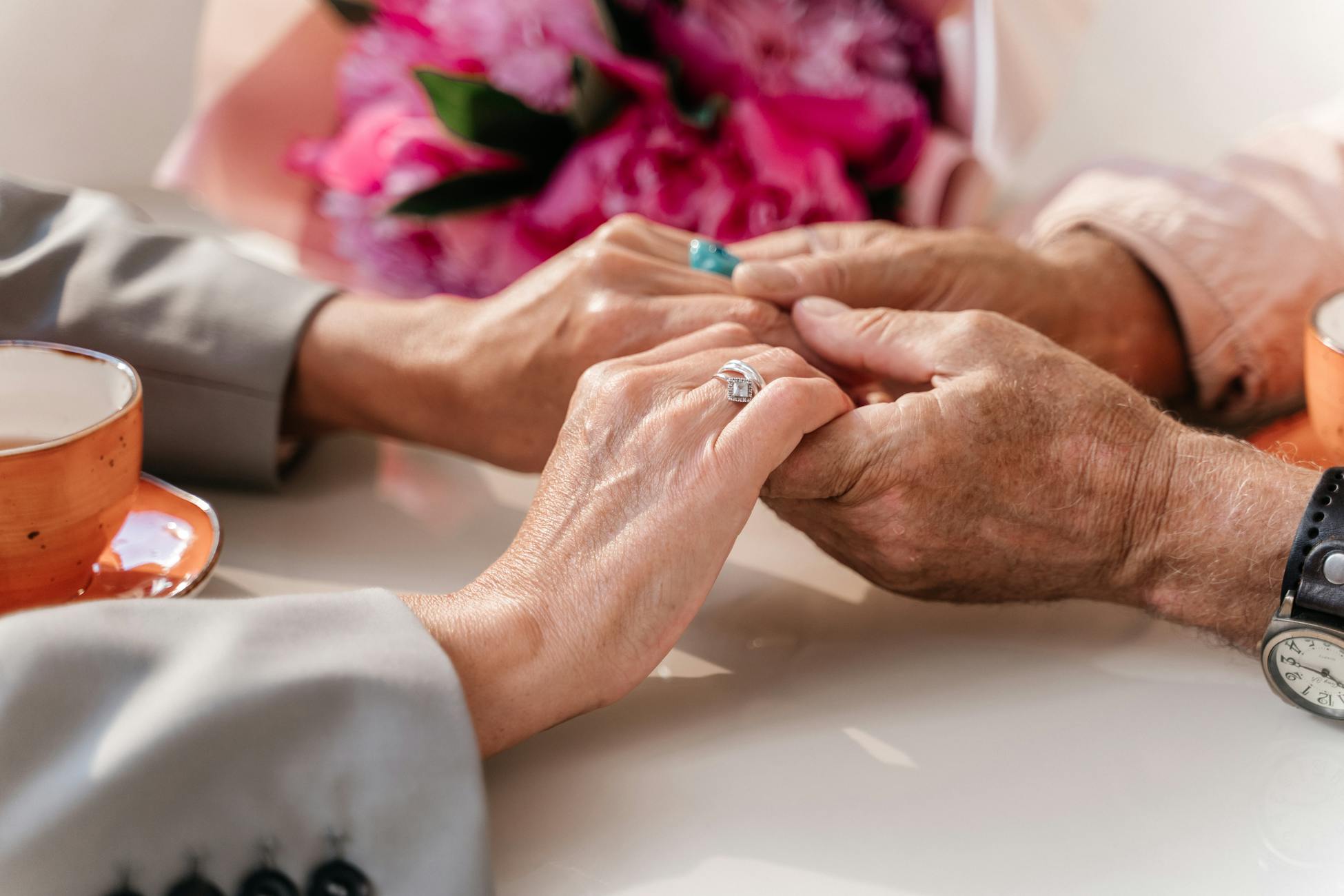 An elderly couple shares a tender moment, holding hands over a table with coffee and flowers.