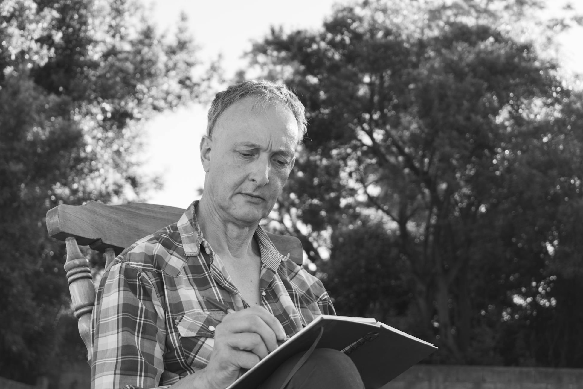 An elderly man in a plaid shirt sits outdoors writing in a notebook, captured in black and white.