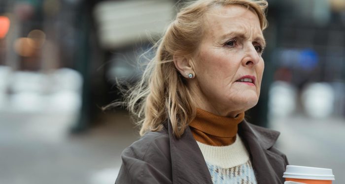 Pensive elderly female with takeaway hot drink looking away in town on windy day on blurred background