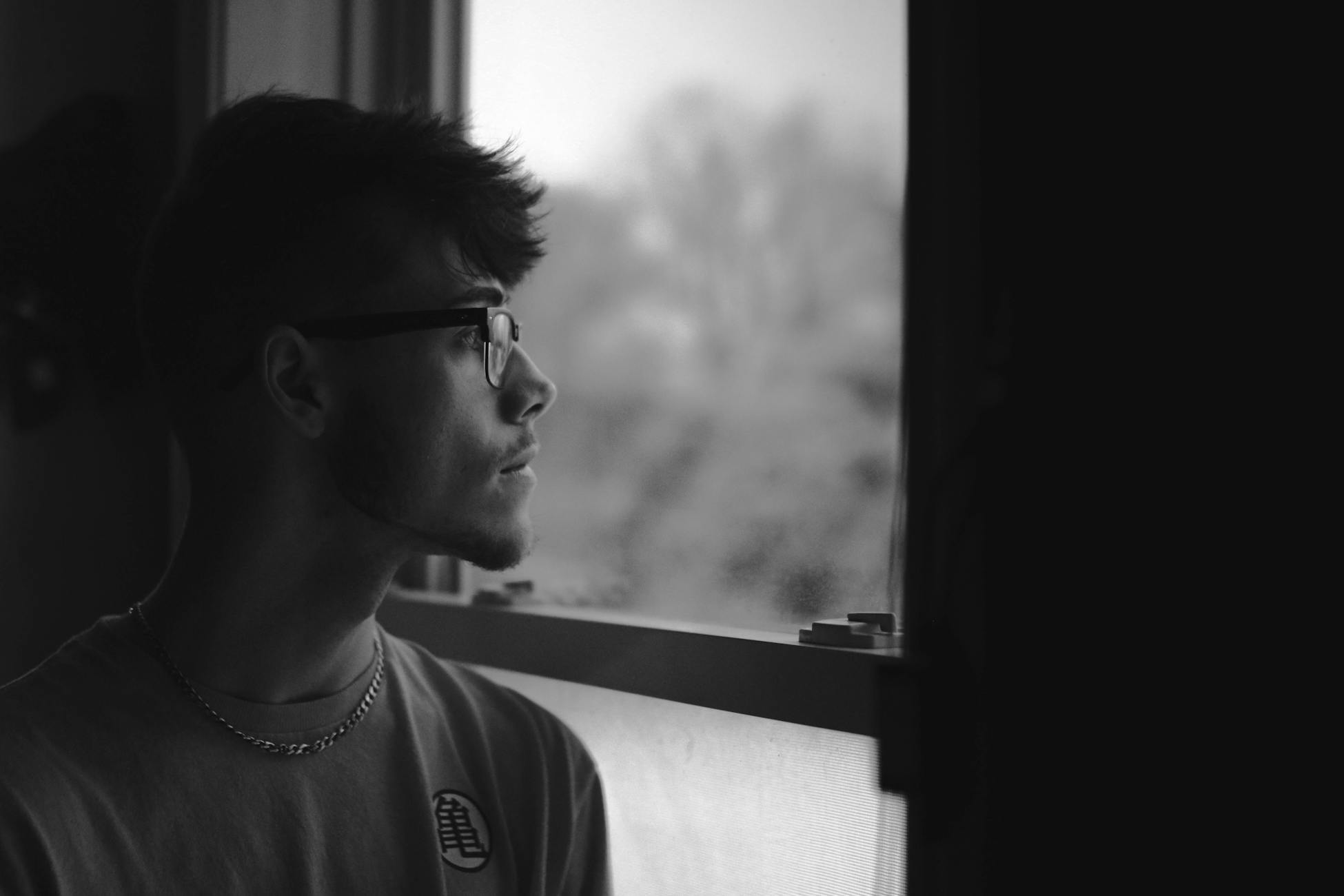 Black and white portrait of a man in eyeglasses looking out a window, feeling pensive.