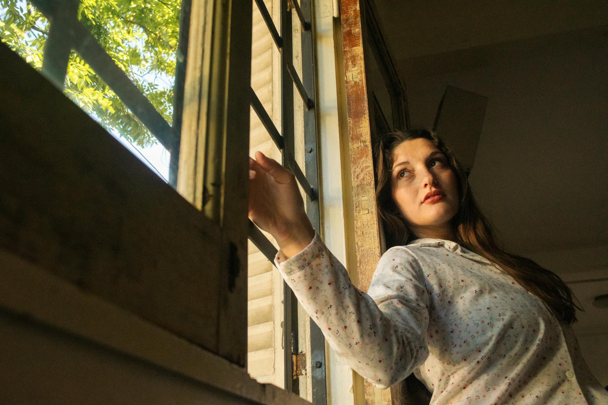 A thoughtful woman stands by an open window, bathed in warm sunlight, indoors.