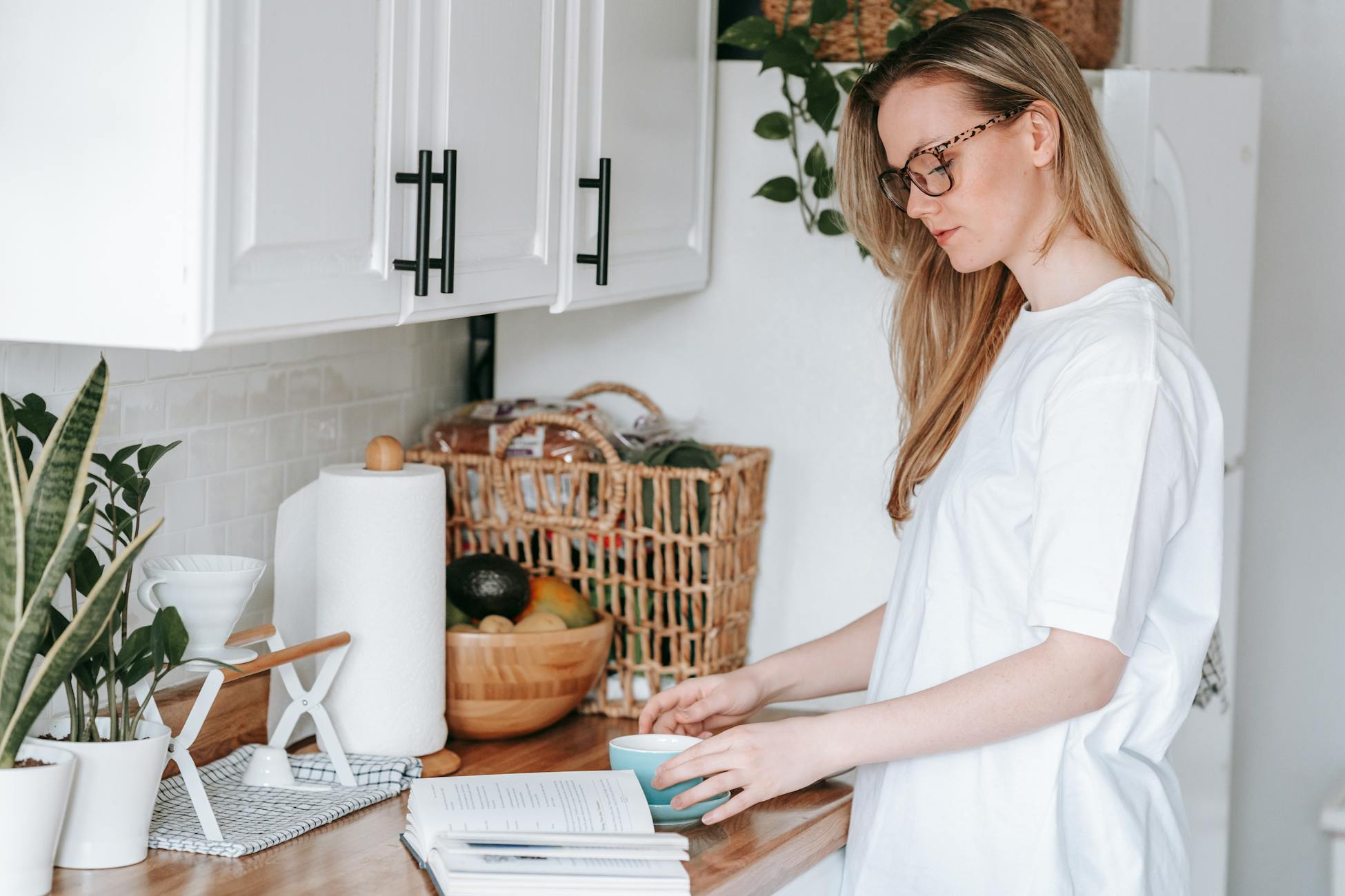 Side view of young female in eyeglasses reading book at wooden counter with mug of hot beverage in light kitchen