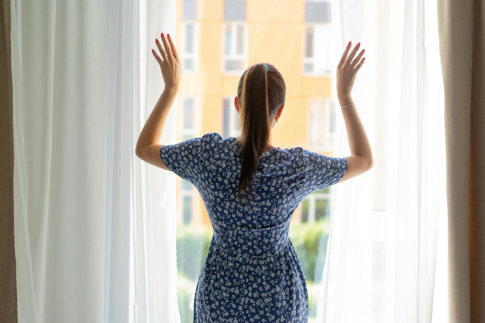 A woman in a floral dress stands looking out through light curtains in a bright room.
