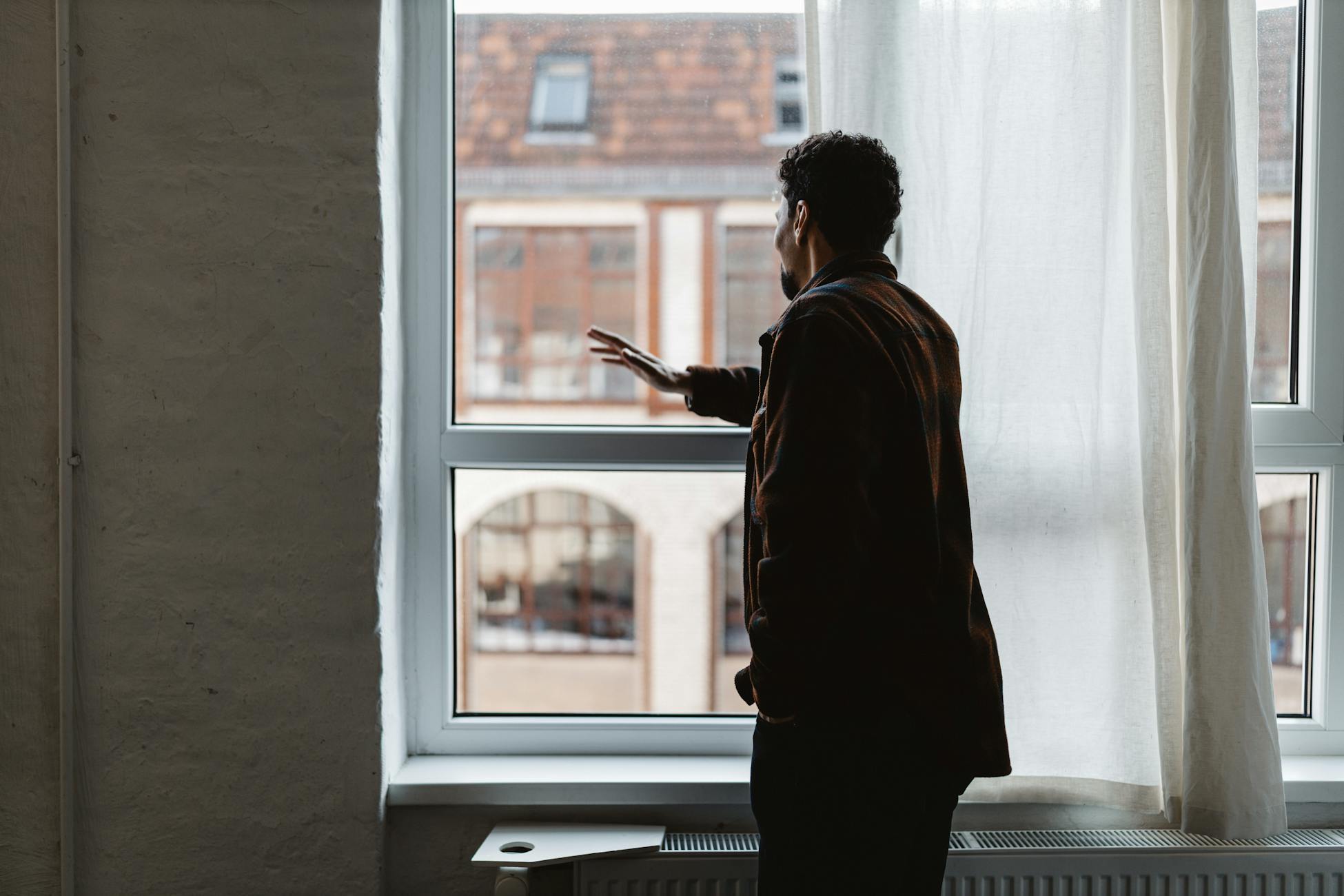 An adult gazing thoughtfully out a large window in a softly lit room.