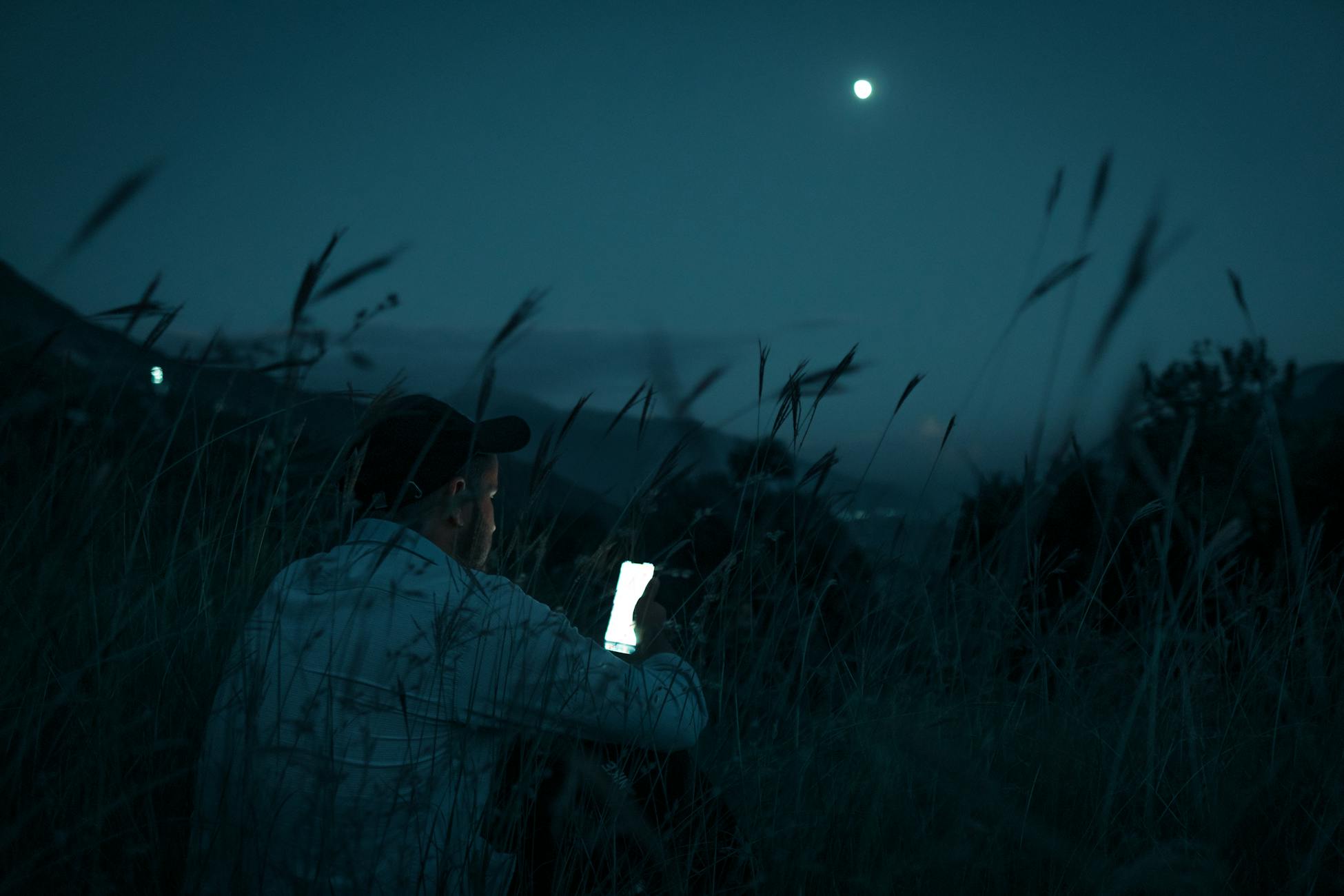 A man sits in a grassy field using his smartphone under the moonlight.