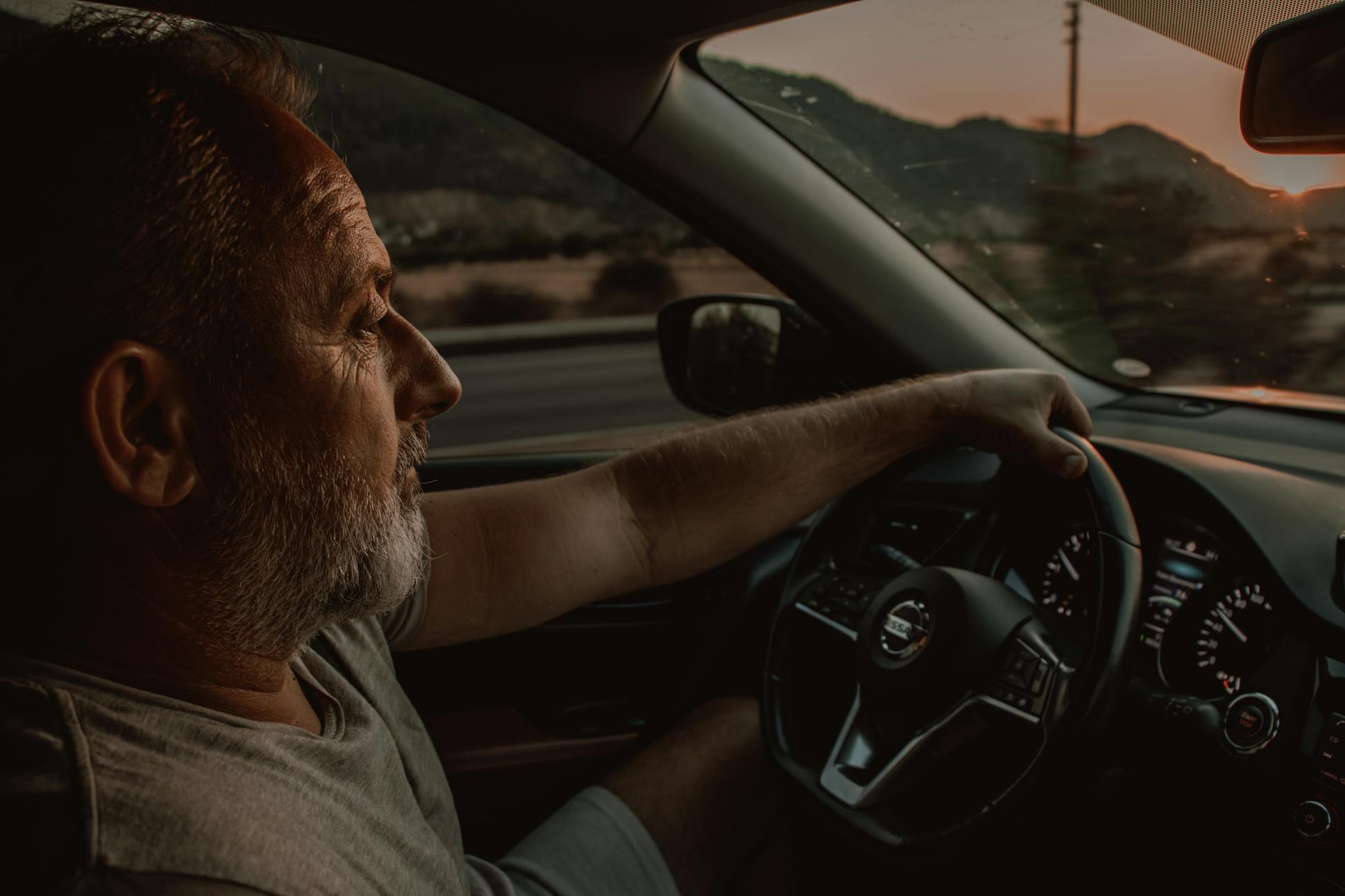 Elderly man driving a Nissan car during sunset, depicting travel and contemplation.