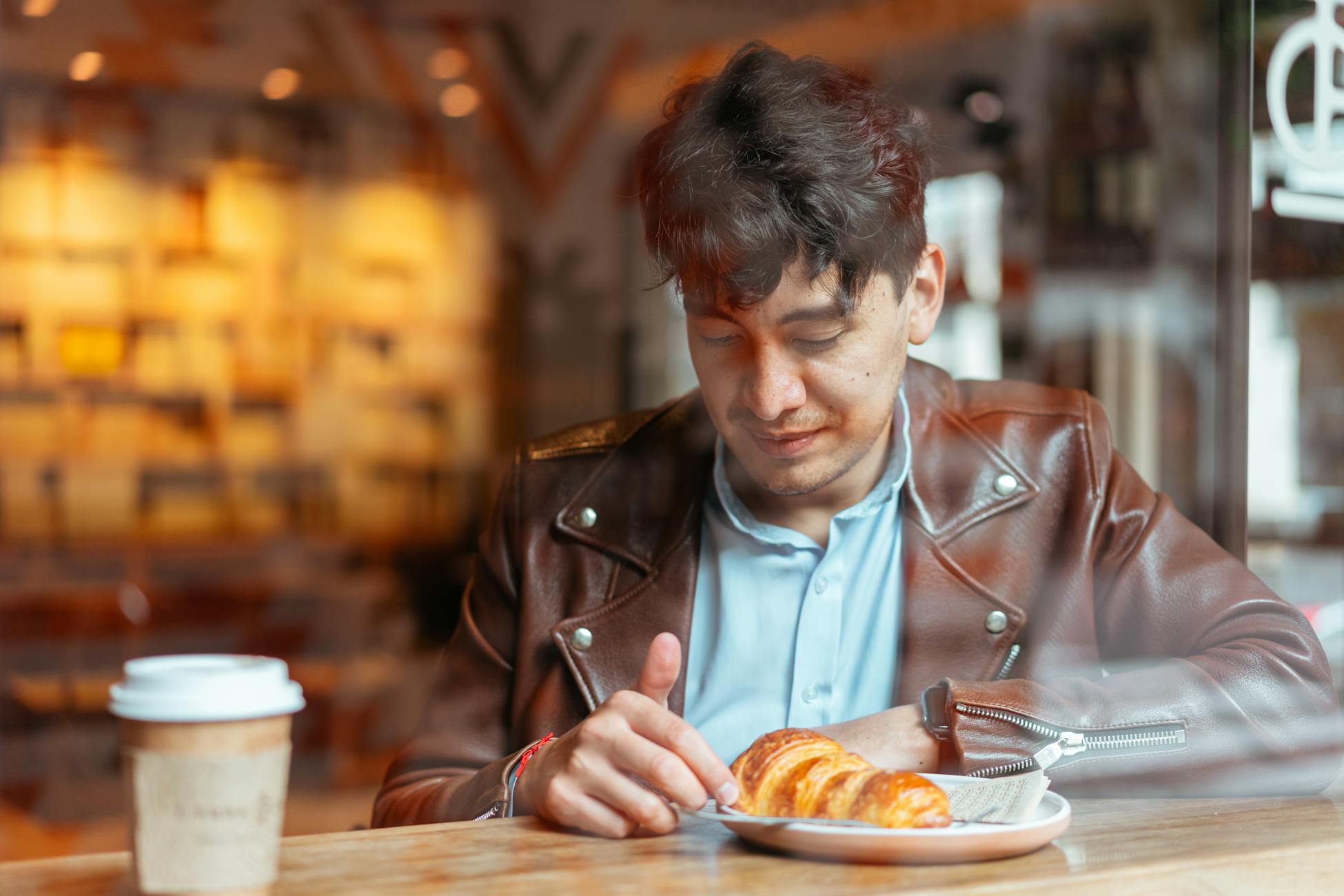 A man in a coffee shop enjoys a croissant and coffee, seen through glass.