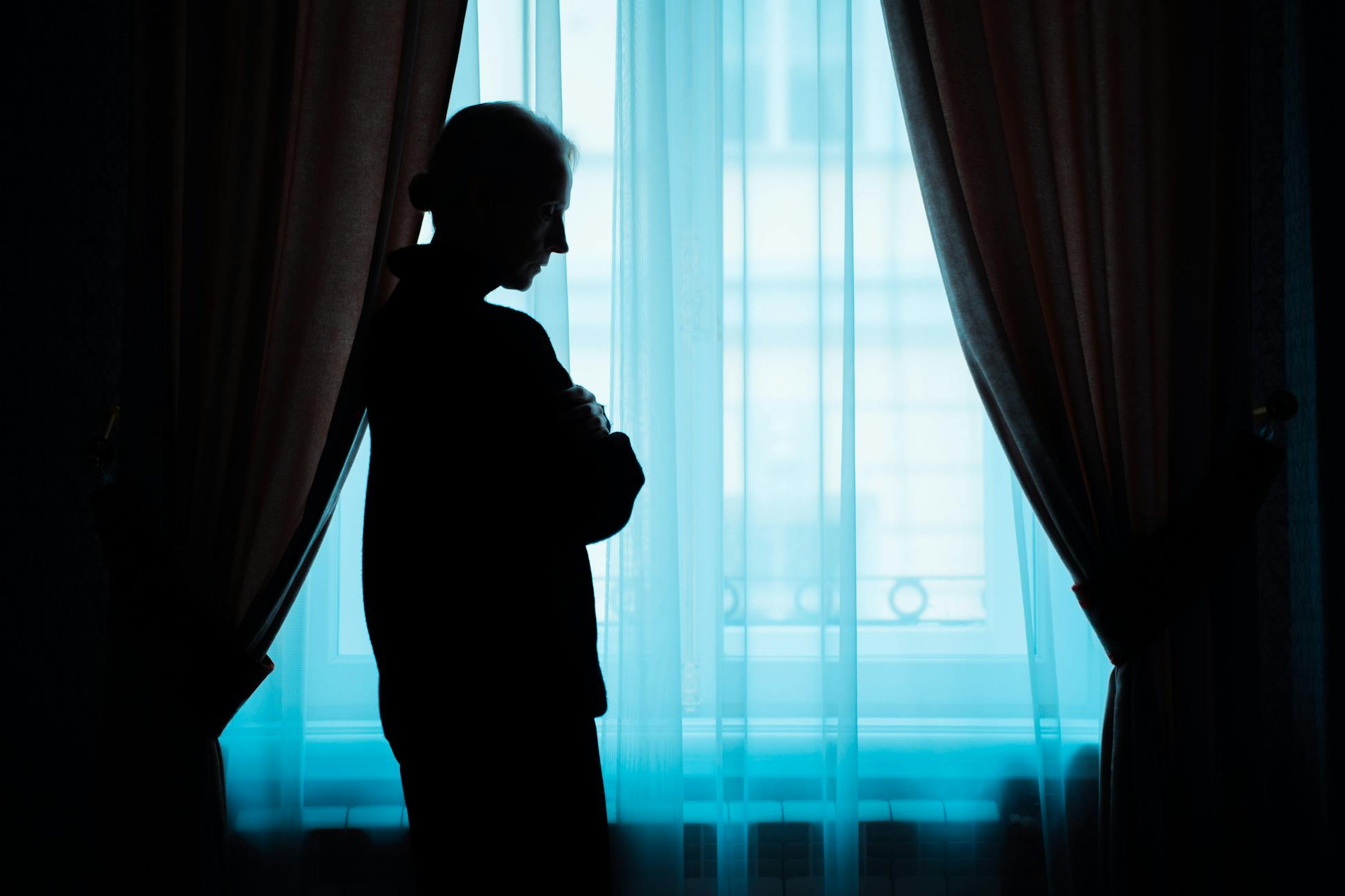 A silhouette of a senior woman stands thoughtfully by a window, conveying solitude.