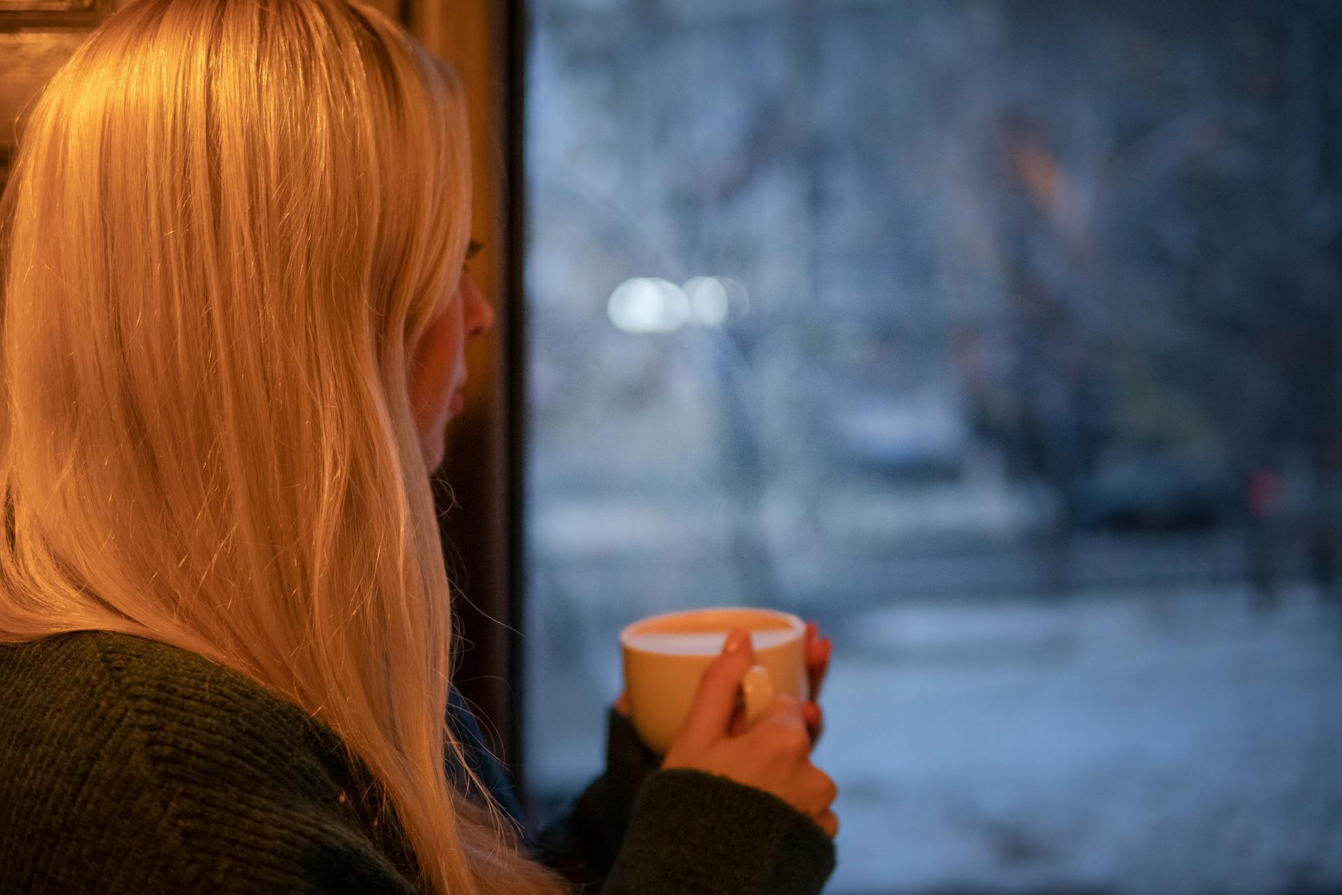 A woman with blonde hair relaxes indoors with a cup of coffee, looking out at the winter scene.